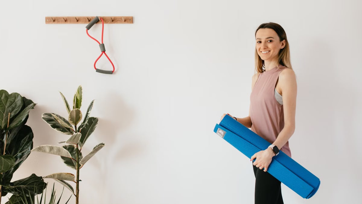 Fit female athlete in sportswear standing against white wall near potted plants and expander on wall hanger while carrying blue fitness mat and looking at camera with smile